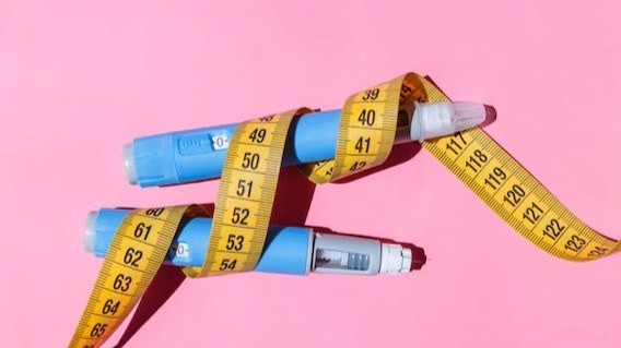 Two blue insulin pens wrapped with a yellow measuring tape against a pink background